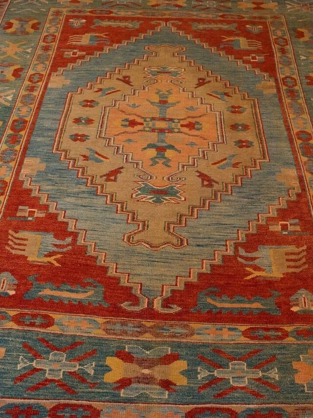 Teppich mit gestuftem Medaillon in Sand und Hellblau auf rotem Grund, umrahmt von einer türkisfarbenen Bordüre